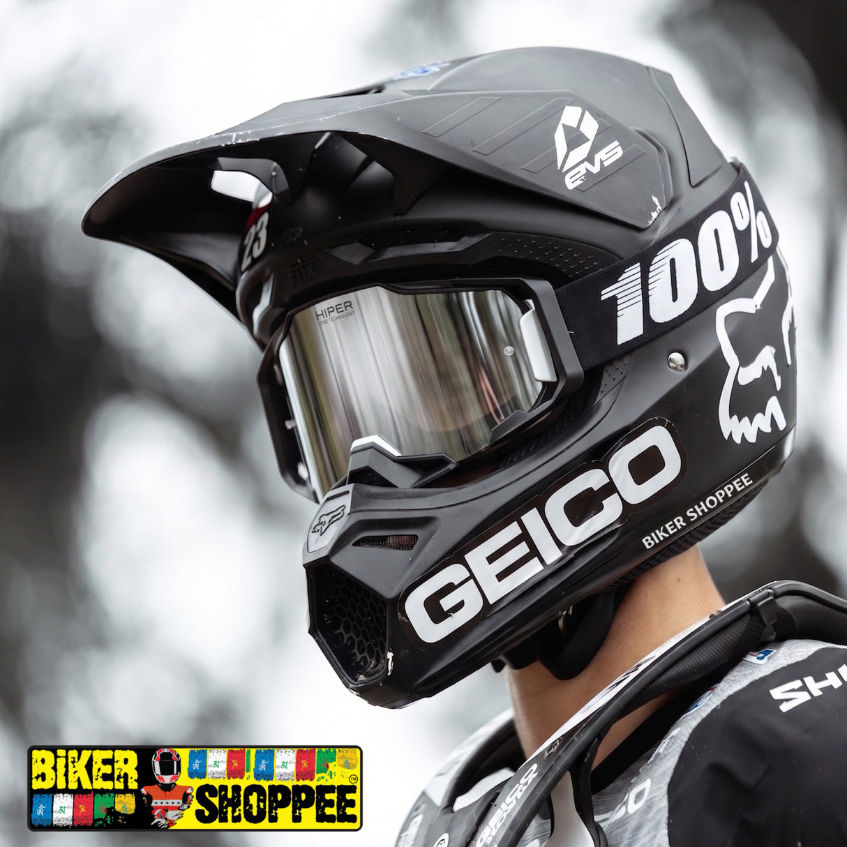 A close-up of a black full-face motocross helmet with a clear visor, worn by a rider. The helmet features prominent white branding for 'GEICO' and '100%' along with the Fox Racing fox head logo.