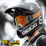 A close-up of a black full-face mountain bike helmet with a reflective orange-tinted visor, worn by a rider. The helmet features prominent white and orange graphics, including the '100%' text and the Fox Racing fox head logo.
